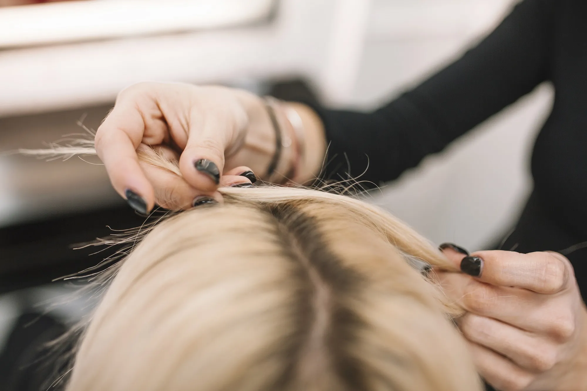 Close-up van handen die lokken blond haar bij de scheiding uit elkaar houden, waarbij de donkere uitgroei zichtbaar is.